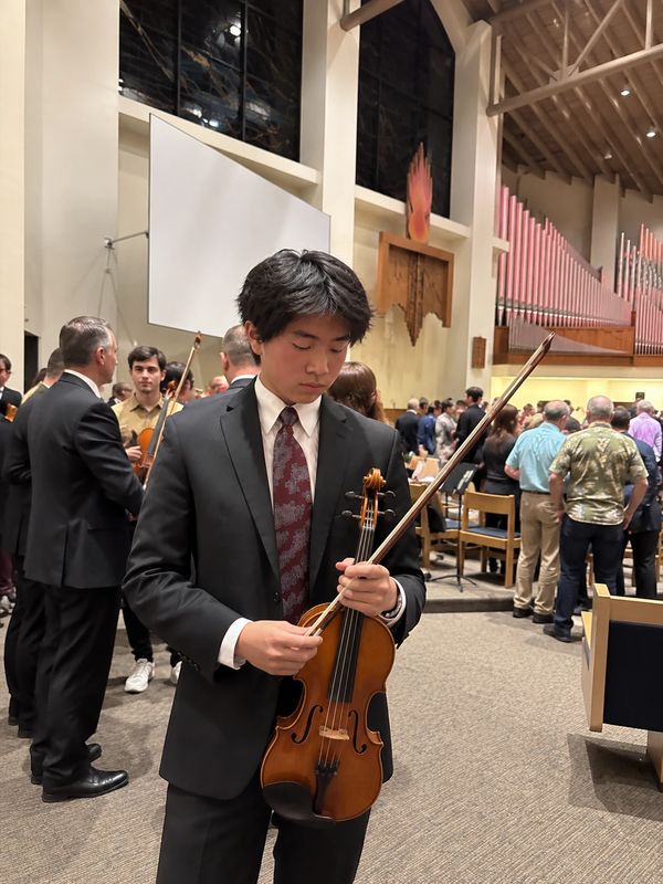 Jonathan in a dark suit and patterned tie holds his violin and bow after a concert. He stands in a hall with organ pipes, with other musicians and the audience behind him.